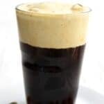 Titled image of a glass of Keto Pistachio Cream Cold Brew on a white plate on a white wooden table.