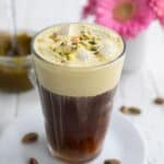 A glass filled with pistachio cream cold brew on a white table with a vase of pink flowers in the background.