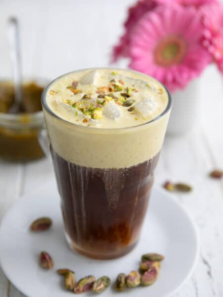 A glass filled with pistachio cream cold brew on a white table with a vase of pink flowers in the background.