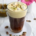 A glass filled with pistachio cream cold brew on a white table with a vase of pink flowers in the background.