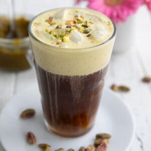 A glass filled with pistachio cream cold brew on a white table with a vase of pink flowers in the background.