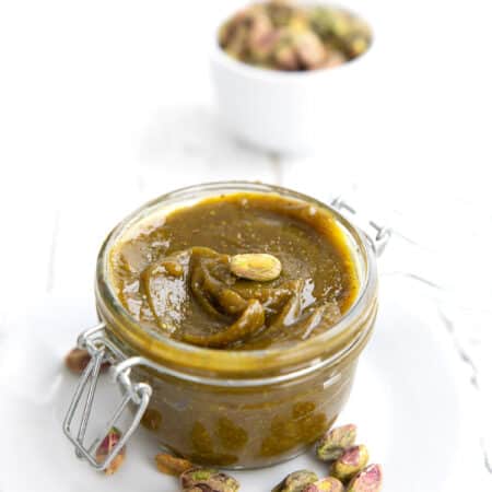 A glass jar filled with keto pistachio cream on a white plate, with a bowl of pistachios in the background.