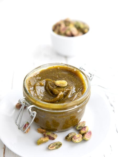 A glass jar filled with keto pistachio cream on a white plate, with a bowl of pistachios in the background.