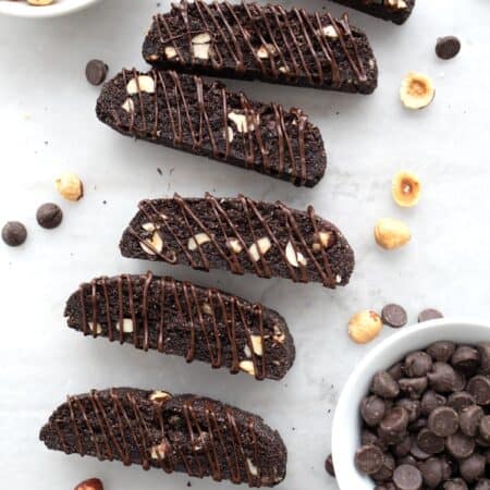 Top down image of keto chocolate hazelnut biscotti laid on their sides with chocolate drizzled over and a bowl of hazelnuts and chocolate chips.