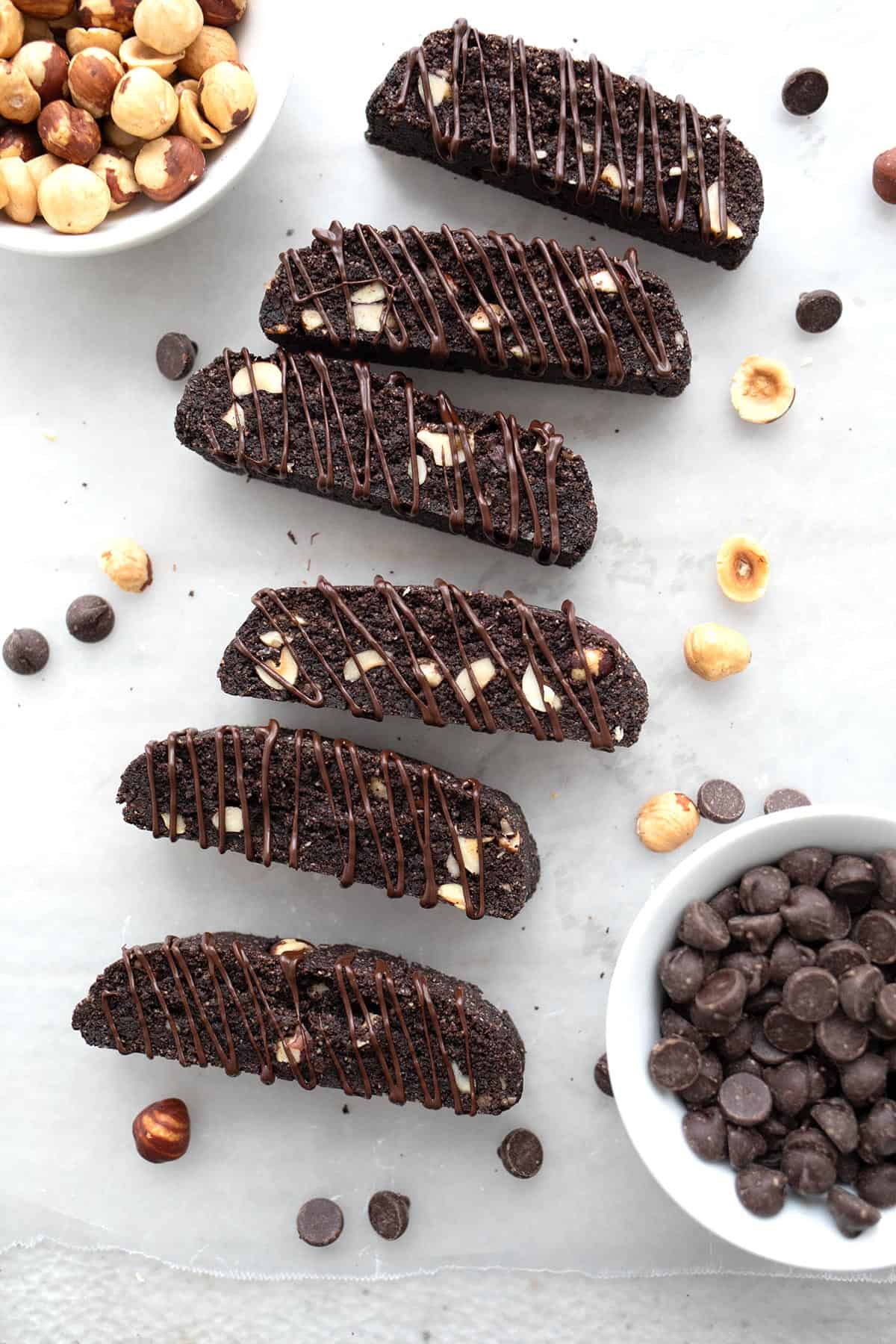 Top down image of keto chocolate hazelnut biscotti laid on their sides with chocolate drizzled over and a bowl of hazelnuts and chocolate chips.