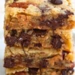 Titled image of a stack of Loaded Keto Cookie Bars on a white plate, with pecans and chocolate chips around the base.