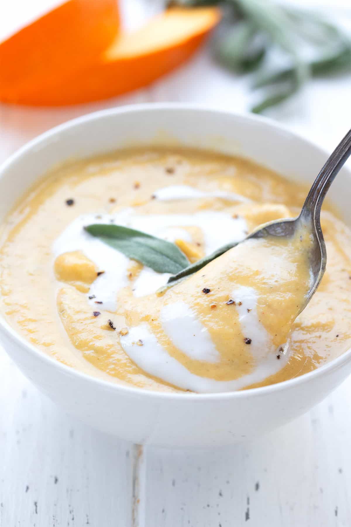 A spoon digging into a white bowl of Creamy Keto Pumpkin Soup, on a white wooden table.