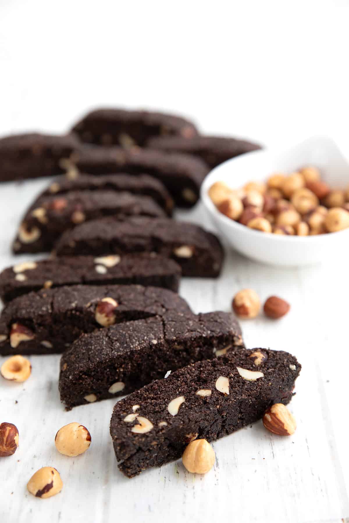 Keto Chocolate Hazelnut Biscotti sliced, in a row on a white table, with hazelnuts strewn around.