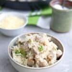 A bowl of chicken and cauliflower rice on a gray table, with the pan filled with more chicken and cauliflower rice in the background.