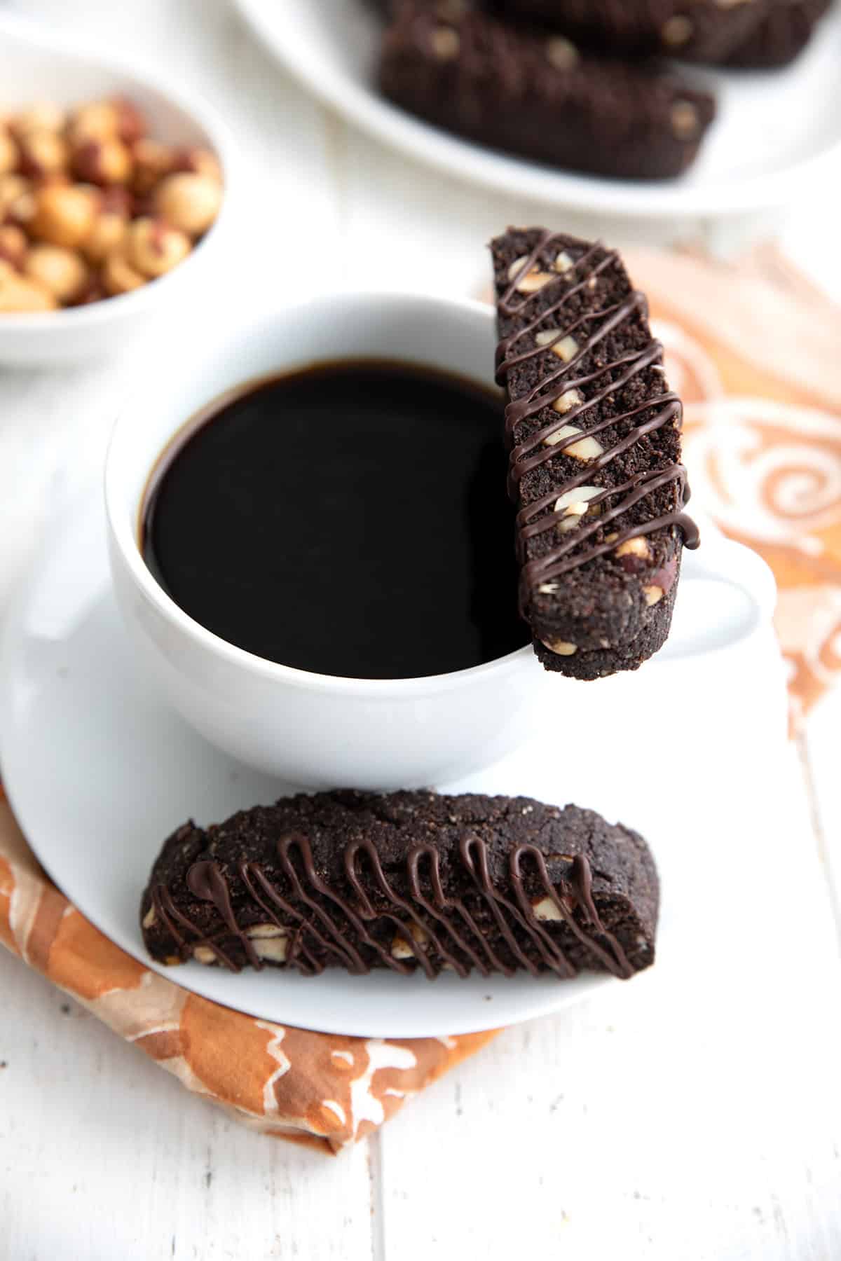 A keto chocolate biscotto resting on the edge of a white cup of coffee, with another biscotto on the saucer.