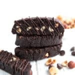 Three chocolate biscotti with chocolate drizzle stacked up on a white wooden table, with hazelnuts and chocolate chips strewn around.