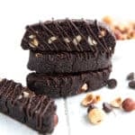 Three chocolate biscotti with chocolate drizzle stacked up on a white wooden table, with hazelnuts and chocolate chips strewn around.