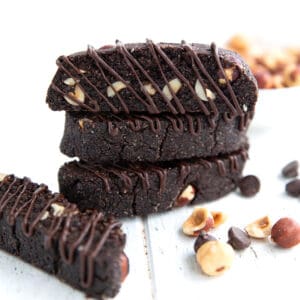 Three chocolate biscotti with chocolate drizzle stacked up on a white wooden table, with hazelnuts and chocolate chips strewn around.