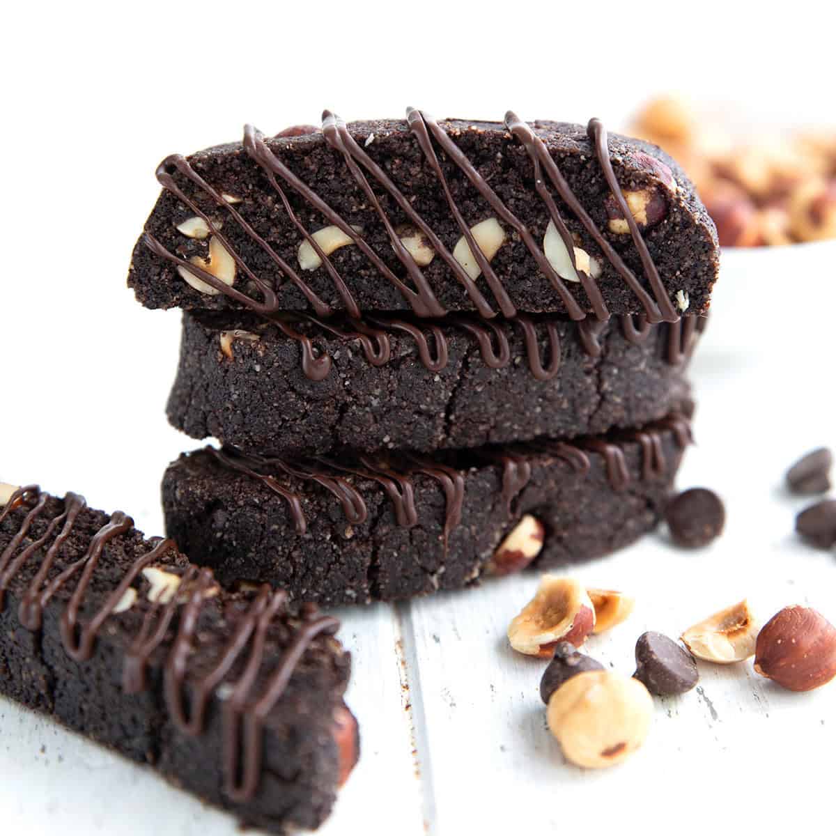 Three chocolate biscotti with chocolate drizzle stacked up on a white wooden table, with hazelnuts and chocolate chips strewn around.