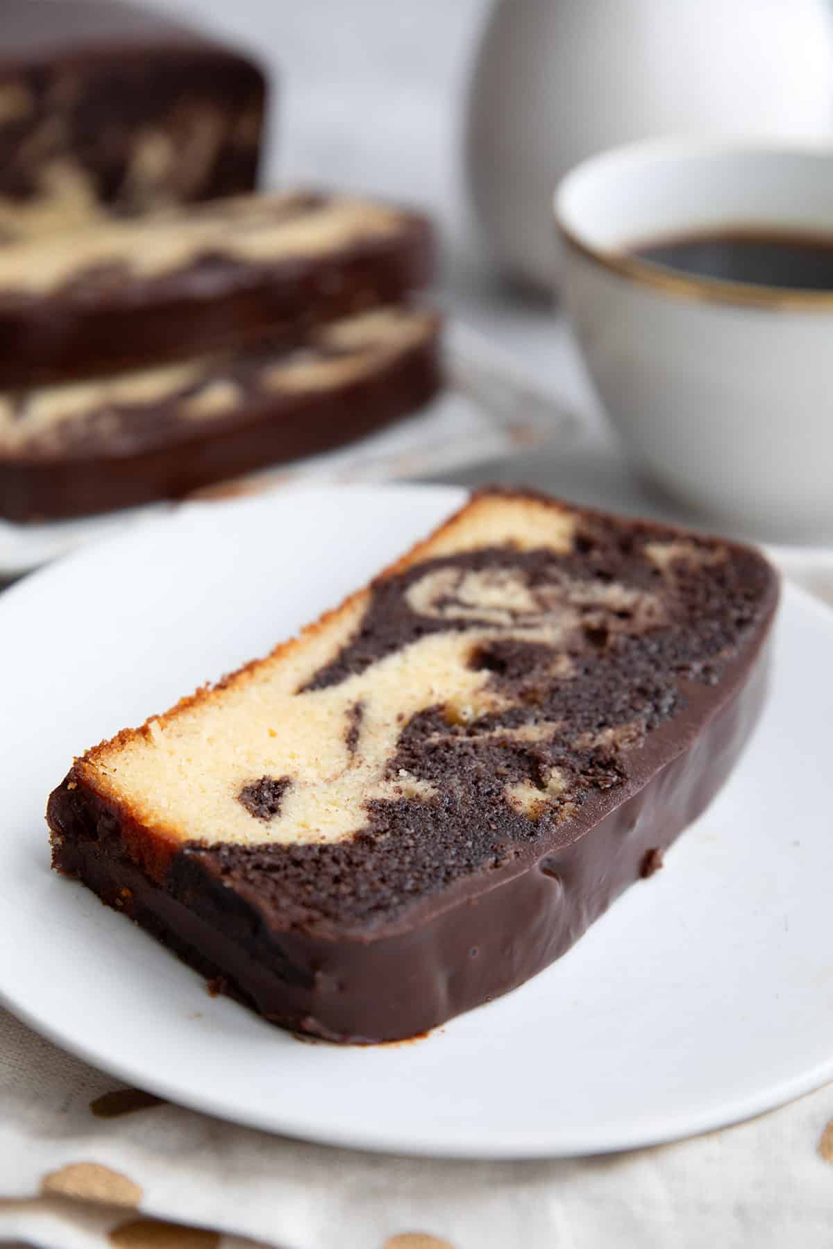 A slice of keto marble pound cake on a white plate with a cup of coffee in the background.