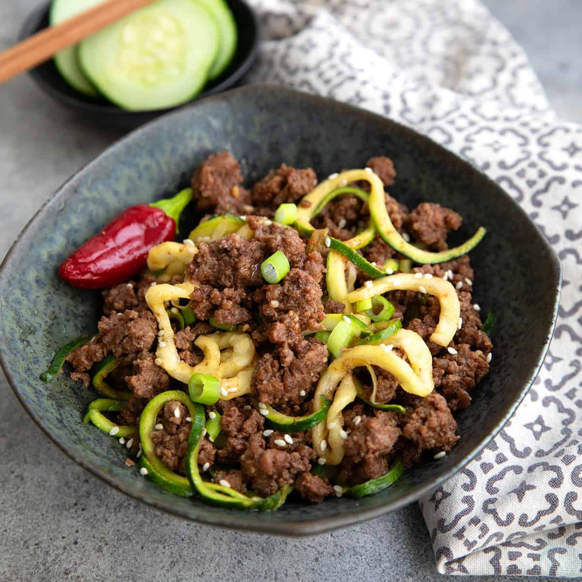 A black bowl filled with Keto Korean Beef and Zucchini Noodles, with a small bowl of cucumbers in the background.