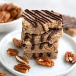 Four keto pecan pie protein bars with chocolate drizzle in a stack on a white plate, with a bite taken out of the top one.