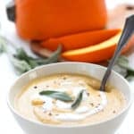 A white bowl filled with keto pumpkin soup on a white wooden table, with a pie pumpkin in the background.