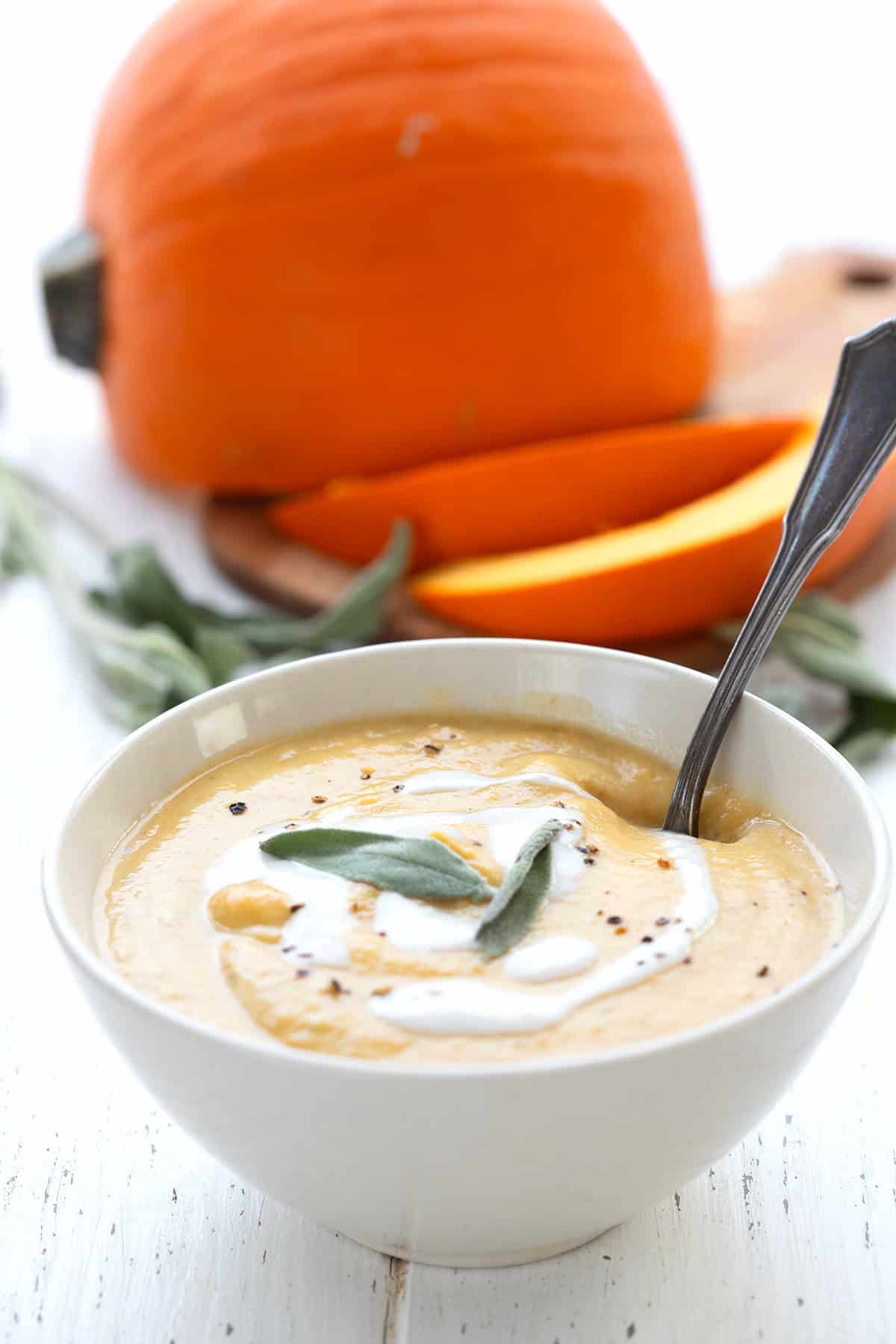 A white bowl filled with keto pumpkin soup on a white wooden table, with a pie pumpkin in the background.