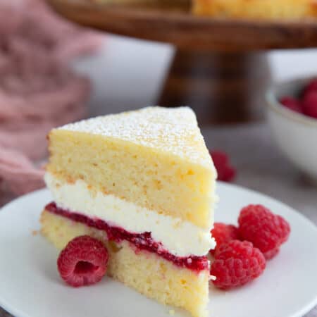 A thick slice of two layer almond flour cake with a raspberry jam layer and a whipped cream layer in the center, on a white plate with fresh raspberries.