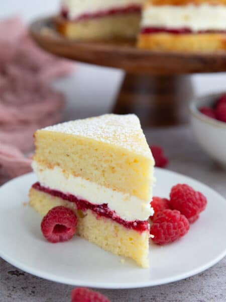 A thick slice of two layer almond flour cake with a raspberry jam layer and a whipped cream layer in the center, on a white plate with fresh raspberries.