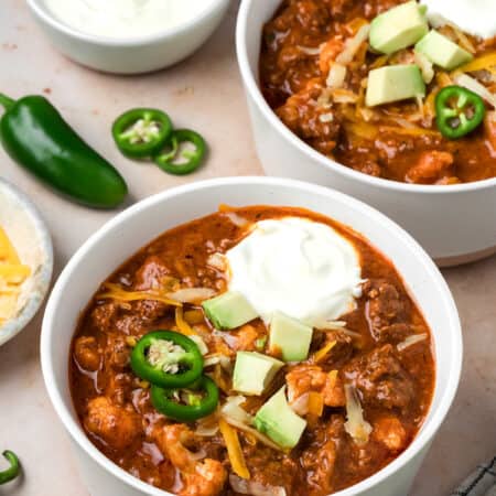 Two white bowls filled with Keto Chili Mac, topped with sliced jalapeño, sour cream, and avocado