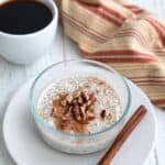 A small glass jar filled with keto overnight oats with chopped pecans and cinnamon on top, with a cup of coffee in the background.