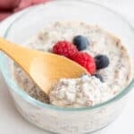 A wooden spoon digging into a glass container filled with overnight keto chia seed cereal with fresh berries on top.