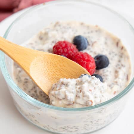 A wooden spoon digging into a glass container filled with overnight keto chia seed cereal with fresh berries on top.