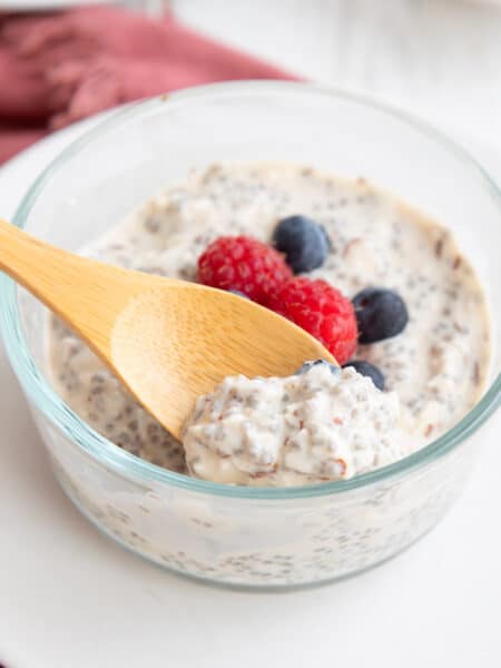 A wooden spoon digging into a glass container filled with overnight keto chia seed cereal with fresh berries on top.