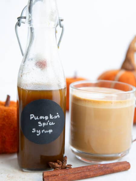 A bottle of Keto Pumpkin Spice Syrup beside a cup of coffee with frothed milk.